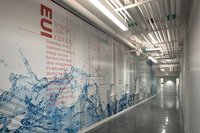 A modern corridor in the Engineering & Interdisciplinary Sciences Complex featuring a large graphic wall explaining energy use intensity, with industrial-style lighting overhead.