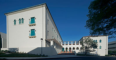 Full view of the Engineering & Interdisciplinary Sciences Complex. A large, white stucco building with a Mediterranean architectural style featuring arched windows with turquoise shutters, decorative grilles, and a terracotta roof.