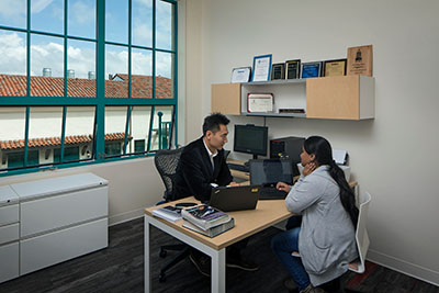 A student and a professor discussing over a laptop in a modern office in the Engineering & Interdisciplinary Sciences Complex with large teal windows overlooking a Mediterranean-style roof.