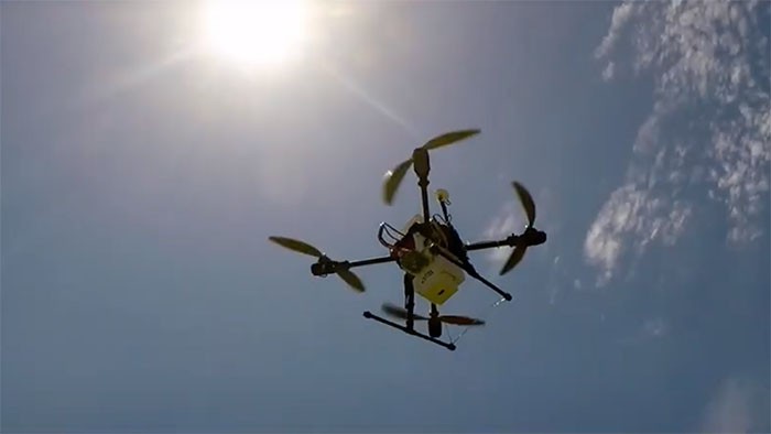 A quadcopter drone flying in a clear blue sky with the bright sun overhead, partially obscured by the drone’s silhouette. The drone has four propellers and is equipped with a small payload attachment beneath its frame.
