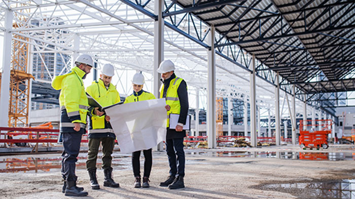 Group of construction and civil engineering professionals in safety gear reviewing blueprints at a large steel-frame building site.